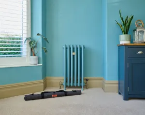 A short Emmeline III cast iron radiator painted to match a turquoise wall, with angled valves, beside a window and a rolled tool kit on the carpet.
