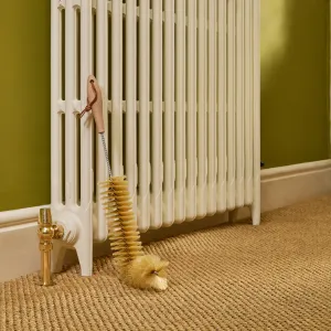Redecker radiator brush with wooden handle hanging on a cream cast iron column radiator, beside a brass valve on a sisal carpet against green walls.