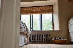 A low Grace 4 Column cast iron radiator in Grey Bronze beneath a bedroom window with a bamboo blind, beside a bed and a wooden bowl.