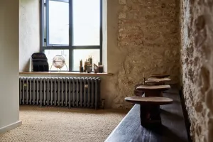 Grace 4 Column cast iron radiator in Grey Bronze on feet beneath a window, in a stone-walled room with sisal carpet and a wooden bench.