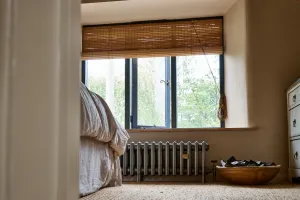 Low Grace 4 column cast iron radiator in Grey Bronze on feet beneath a bedroom window with bamboo blind, beside a bed and wooden bowl.