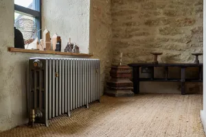 Grace 4 Column cast iron radiator in Grey Bronze on feet with a brass valve, below a window sill in a stone-walled room with jute carpet.