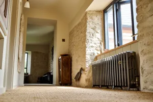 Grace 4 Column 500mm cast iron radiator in Grey Bronze on feet beneath a window, beside exposed stone walls and a sisal-carpeted landing.