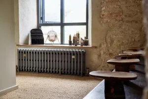 Grace 4 Column cast iron radiator in Grey Bronze on feet beneath a window, beside a stone wall and wooden stools on a sisal floor.