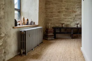 Grace 4 Column cast iron radiator in Grey Bronze on feet with brass valves, under a window in a stone-walled room with sisal flooring.