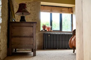 Grace 4 Column cast iron radiator in Grey Bronze under a window in a stone-walled bedroom, beside an oak chest and lamp on sisal carpet.