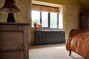 Grace 4 Column 660mm cast iron radiator in Grey Bronze on feet beneath a window in a stone-walled bedroom with sisal carpet.
