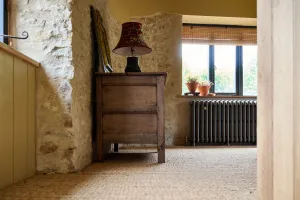 Grace 4 column cast iron radiator in Grey Bronze under a window on feet, beside an oak chest and lamp against a stone wall, sisal carpet.