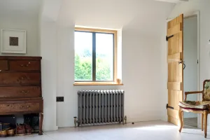 Grace 4 Column cast iron radiator in Grey Bronze on feet beneath an oak-framed window, with brass valves, an oak chest and a ledged door.