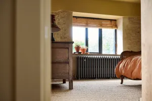 Grace 4 Column cast iron radiator in Grey Bronze under a bedroom window in stone walls, beside an oak chest and a cane bed.