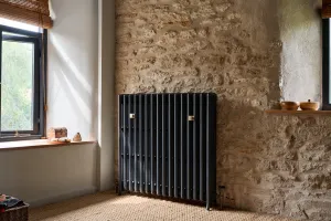 Grace 4 Column 960mm cast iron radiator in Midnight Ink on feet against a stone wall, beside a window, on a seagrass floor at Rodden Mill.