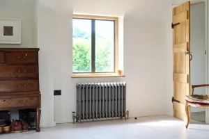 Grace 4 Column cast iron radiator in Grey Bronze on feet beneath a window, with brass valves, an oak chest and plank door nearby.