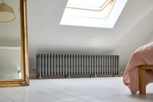 Long Grace 6 Column cast iron radiator in Grey Bronze on feet with a brass valve, under a skylight in a white-floored attic bedroom.