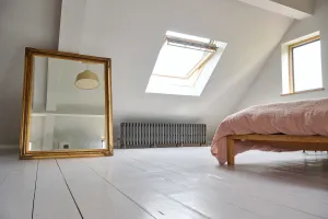 A low Grace 6 Column cast iron radiator in Grey Bronze on feet beneath a roof window in a white attic bedroom with painted floorboards.