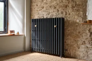 Grace 4 Column 960mm cast iron radiator in Midnight Ink on feet against a stone wall, with a brass valve, beside a window on sisal carpet.