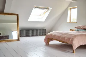 A long, low Grace 6 Column cast iron radiator in Grey Bronze on feet under a skylight in a loft bedroom with white floorboards and a pink bed.