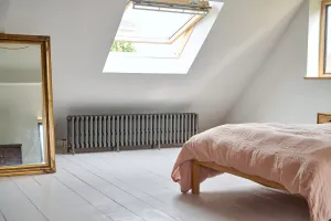 A long, low Grace 6 Column cast iron radiator in Grey Bronze on feet beneath a skylight in a loft bedroom with white floorboards.