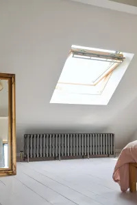 Long, low Grace 6 Column cast iron radiator in Grey Bronze on feet beneath a roof window in a white-floored attic bedroom, with brass valve.