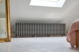 A long, low Grace 6 Column cast iron radiator in Grey Bronze on feet, with a brass valve, beneath a skylight in an attic bedroom with pale floorboards.