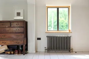 Grace 4 Column cast iron radiator in Grey Bronze with brass valves, under an oak-framed window beside an antique chest of drawers.