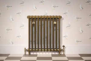 Emmeline I 865mm cast iron radiator in Aged Gold on feet, with brass valves, against seagull wallpaper above a chequered tile floor.