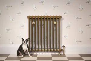 Emmeline I 865mm cast iron radiator in Aged Gold on feet with a Windsor angled valve, seagull wallpaper, chequered floor and a seated dog.