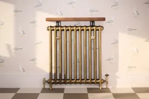 Emmeline I 865mm cast iron radiator in Aged Gold on feet, with Windsor thermostatic valve, wooden shelf above, seagull wallpaper and chequered floor.