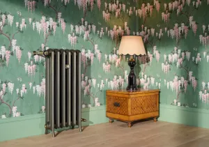 Emmeline II cast iron radiator on feet in a metallic bronze-toned finish with brass valves, beside a marquetry chest and lamp against wisteria wallpaper.