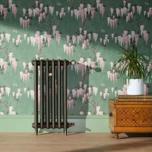 Emmeline II 870mm cast iron radiator in a bronze finish on feet, with brass valves, against green wisteria wallpaper beside a parquetry chest.
