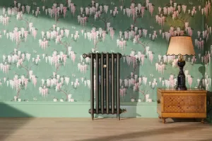 A tall, slim cast iron radiator on feet in a dark bronze-toned finish with a brass valve, set against green wisteria wallpaper beside a lamp on a parquetry chest.