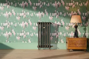 Slim Emmeline II 870mm cast iron radiator in a bronze finish on feet, with brass valves, against green wisteria wallpaper beside a lamp on a chest.