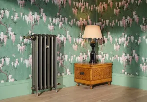 Emmeline II 870mm cast iron radiator on feet in a bronze finish with brass valves, against green wisteria wallpaper beside a parquetry chest.