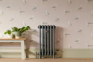 Narrow Grace 4 column cast iron radiator in a dark metallic finish on feet, with Windsor valves, against seagull wallpaper beside a bench.