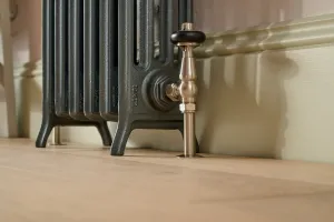 Close-up of a dark grey Grace 4 column cast iron radiator on feet with a Windsor angled thermostatic valve, beside a skirting board on a wooden floor.