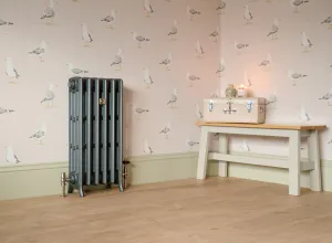 Grace 4 column cast iron radiator in Pencil on feet, with Windsor angled valves, beside a cream bench on a wooden floor against seagull wallpaper.