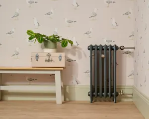 Grace 4 Column 760mm cast iron radiator in a dark grey finish on feet, with a Windsor angled thermostatic valve, beside a bench on a wood floor.