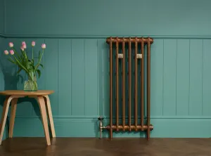 A tall plain Mercury cast iron radiator in a copper finish on feet, with a Windsor angled valve, against teal panelling on a parquet floor.