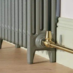 Close-up of the foot end of a Mercury 4 column cast iron radiator in grey-green, with a brass angled valve and pipe by a pale skirting board.