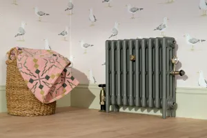 Plain Mercury 4 column cast iron radiator in Quartz Grey on feet with brass valves, beside a wicker basket against seagull wallpaper.