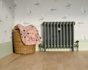 Plain Mercury 4 column cast iron radiator in Quartz Grey on feet with brass Windsor valves, beside a wicker basket on a light oak floor.