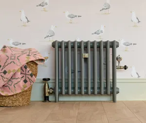 Plain Mercury 4 column cast iron radiator in Quartz Grey on feet, with brass Windsor valves, against seagull wallpaper beside a wicker basket.