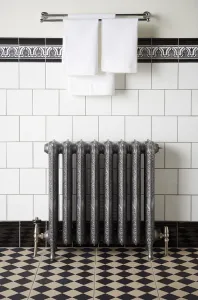Ornate Rococo II cast iron radiator in Etch-Coated Polish on feet with angled valves, below a towel rail on white tiles above a chequered floor.