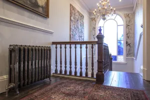 Ornate Rococo III cast iron radiator in Full Polish on feet, with angled valve, on a landing with timber banister, rug and arched window.