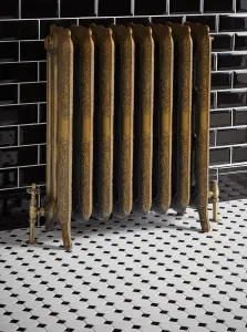 Ornate Rococo III cast iron radiator in Natural Brass on feet, with brass valves, against black metro tiles on a black and white mosaic floor.