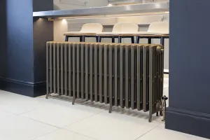 Grace 6 column cast iron radiator in a bronze-toned metallic finish on feet, with a thermostatic valve, beside a kitchen island with bar stools.