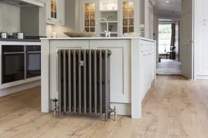 Mercury 4 Column 760mm cast iron radiator in Beaten Brass on feet against a pale kitchen island, with angled valves on an oak floor.