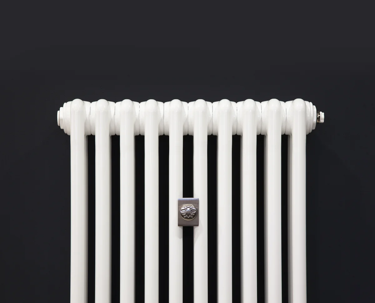 Top of a white Florence 3 Column steel radiator with ten plain tubular sections and a brushed metal bee badge, against a charcoal wall.