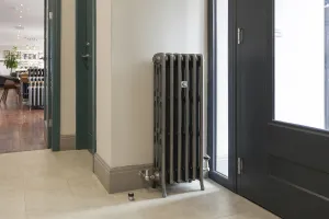 Ornate Grace 4 Column 960mm cast iron radiator in a metallic pewter finish on feet with angled valves, beside a glazed door in the showroom.