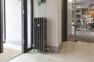 A tall, narrow ornate Grace 4 Column cast iron radiator in a metallic pewter finish on feet, with chrome valves, beside a glass office entrance.