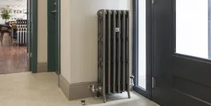 Tall Grace four-column cast iron radiator on feet in a dark metallic finish with nickel valves, beside a glazed door in the showroom.